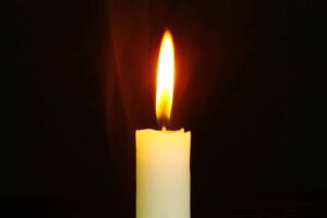 A lit candle with a bright flame against a dark background, with faint wisps of smoke rising from the wick.
