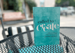 A book titled "Called to Create" by Jordan Raynor displayed on a round metal table. The cover features a blue-green background with white text. The background shows a blurred café setting.