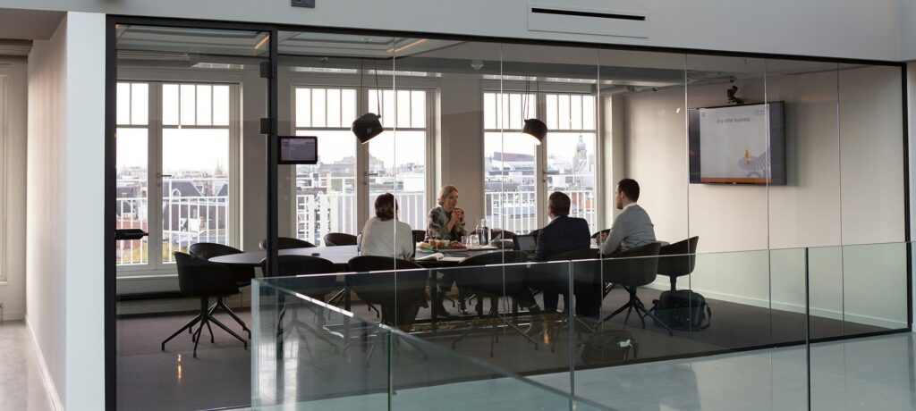 A meeting room with glass walls showing four people around a table, engaged in discussion. A large window provides a view of a city skyline, and a screen displays information on the wall.