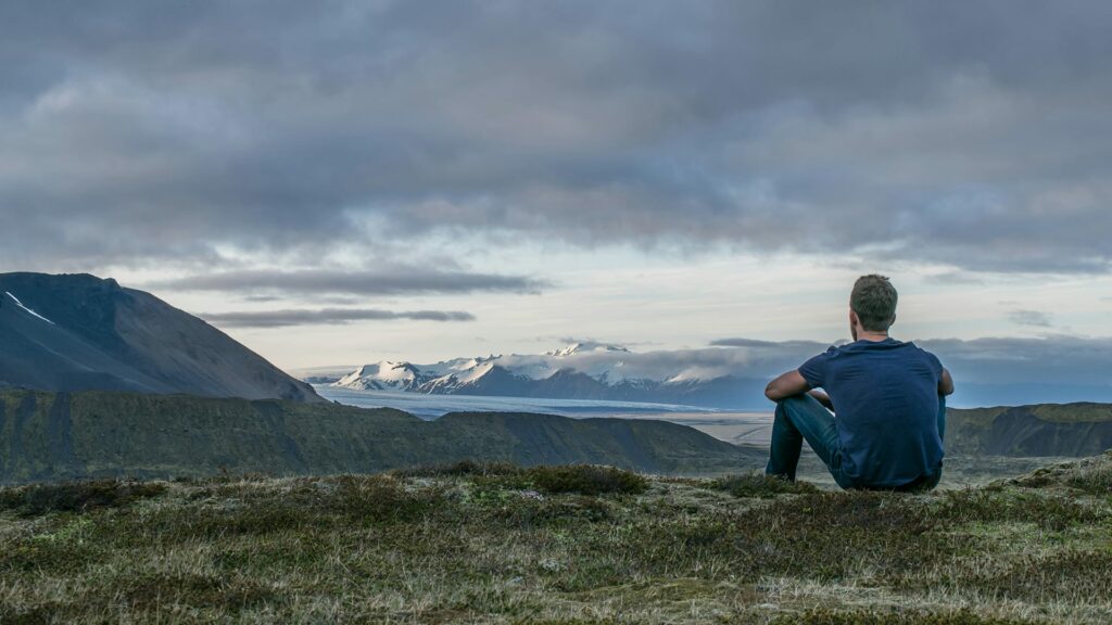 A person sitting on a grassy hill, looking at a mountain range in the distance under a cloudy sky. The landscape features rolling hills and snow-capped peaks.